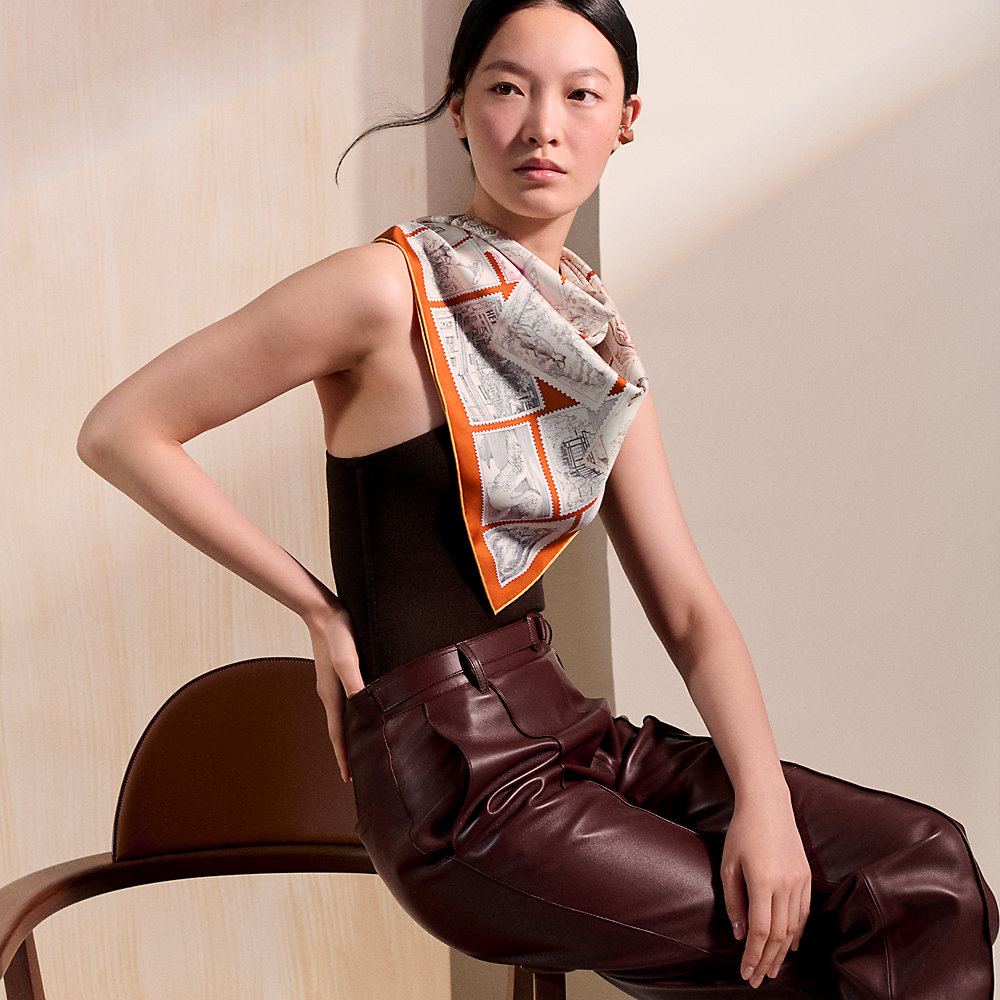 Model wearing an orange Hermès Carré 90 scarf with a black top and burgundy trousers.